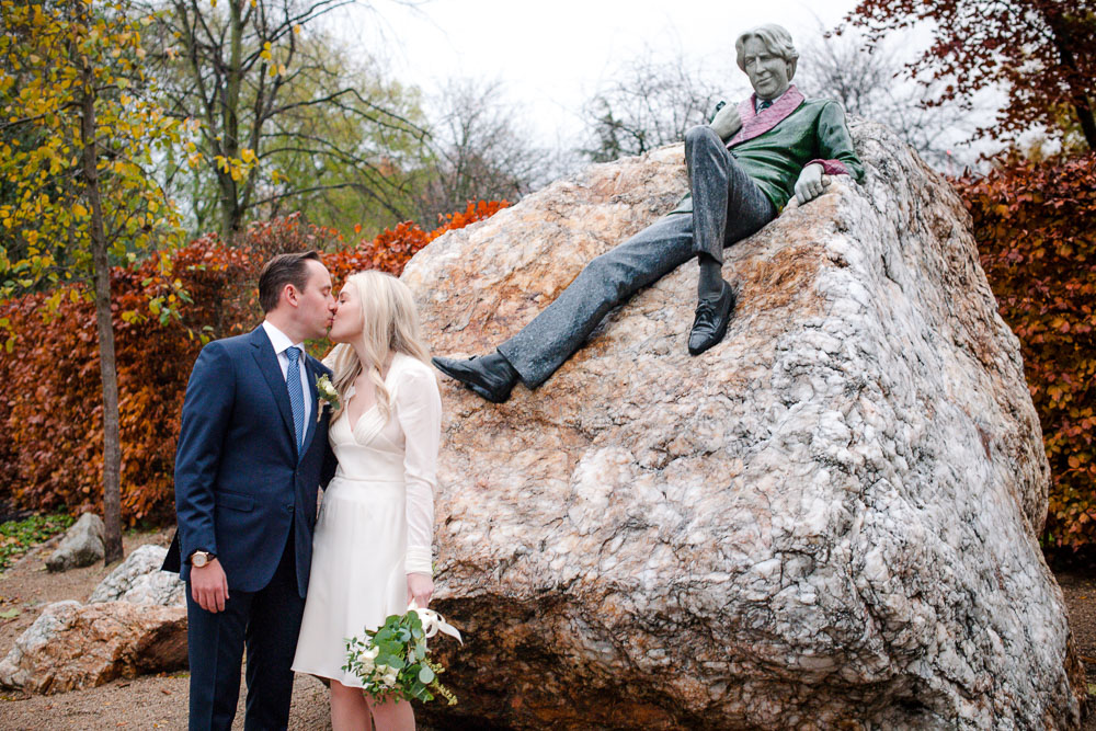 The bride and groom exchange a kiss under the watchful eye of Oscar Wilde in Merrion Square Park in a Merrion Hotel Wedding Photograph
